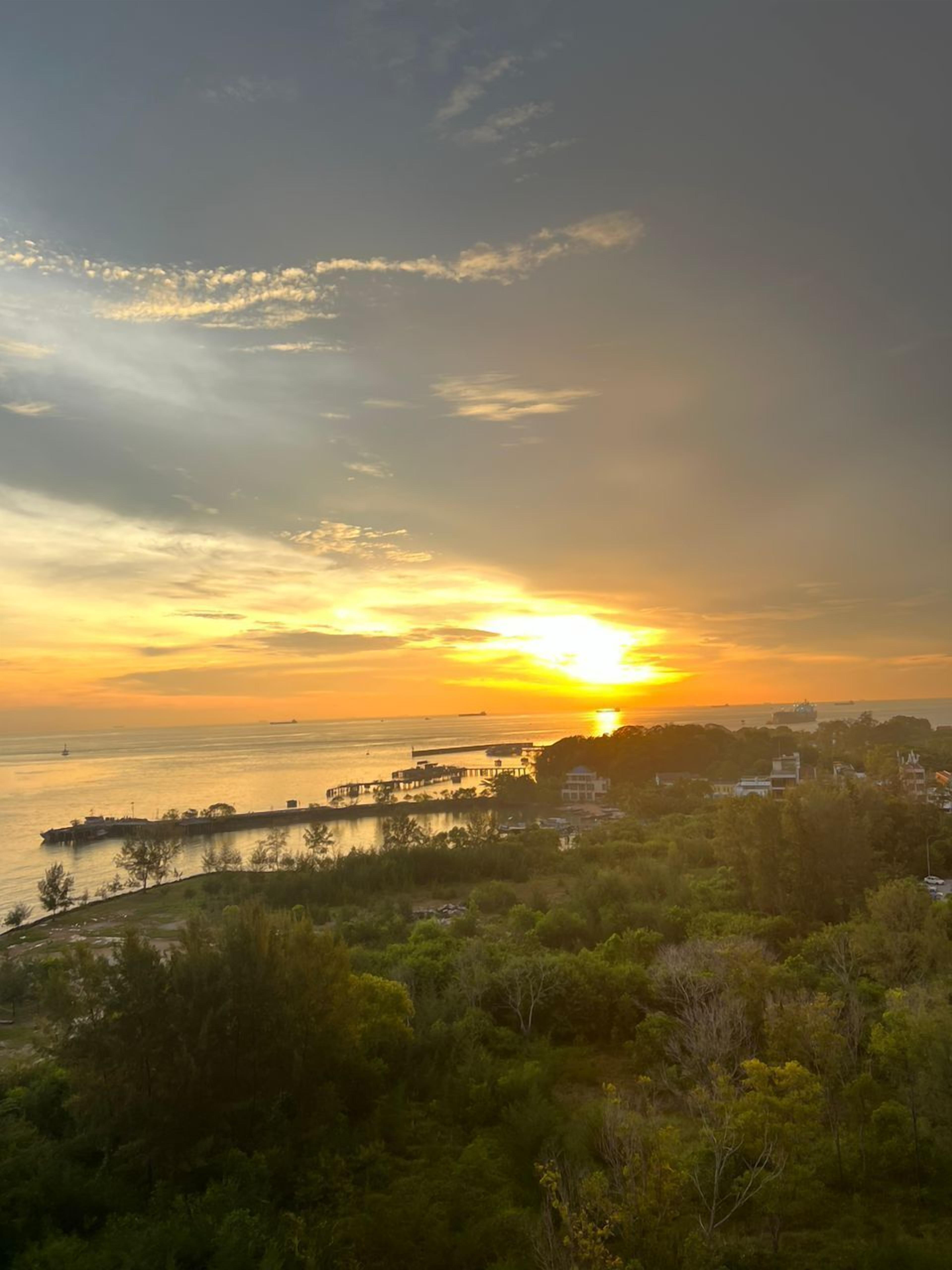 Vista de un atardecer costero con un muelle que se adentra en el agua, rodeado de vegetación y edificios.