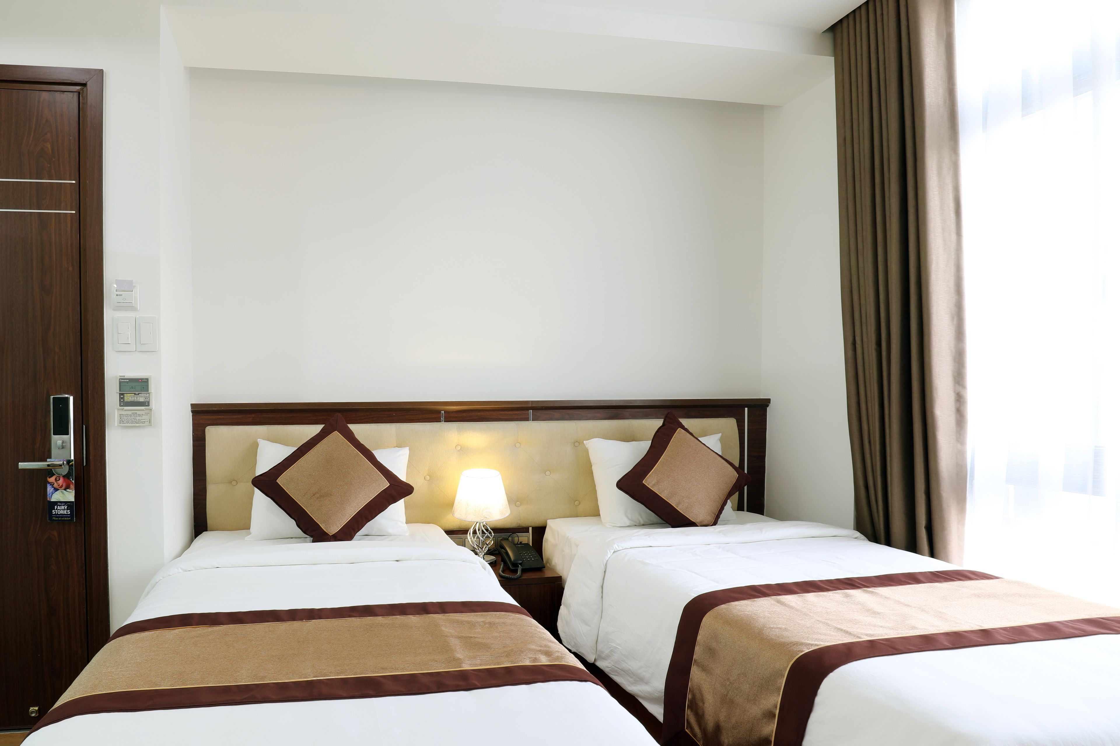 A hotel room with two beds, a wooden headboard, a nightstand with a lamp, and a window with curtains.