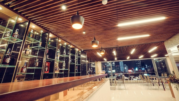 Un bar moderno con una barra alargada, estantes de vidrio con botellas y vista a la ciudad a través de amplias ventanas.