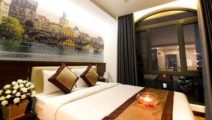 Habitación de hotel con una cama grande, una mesita de noche con un jarrón de flores, una ventana con vistas a un paisaje urbano y un cuadro de un puente.