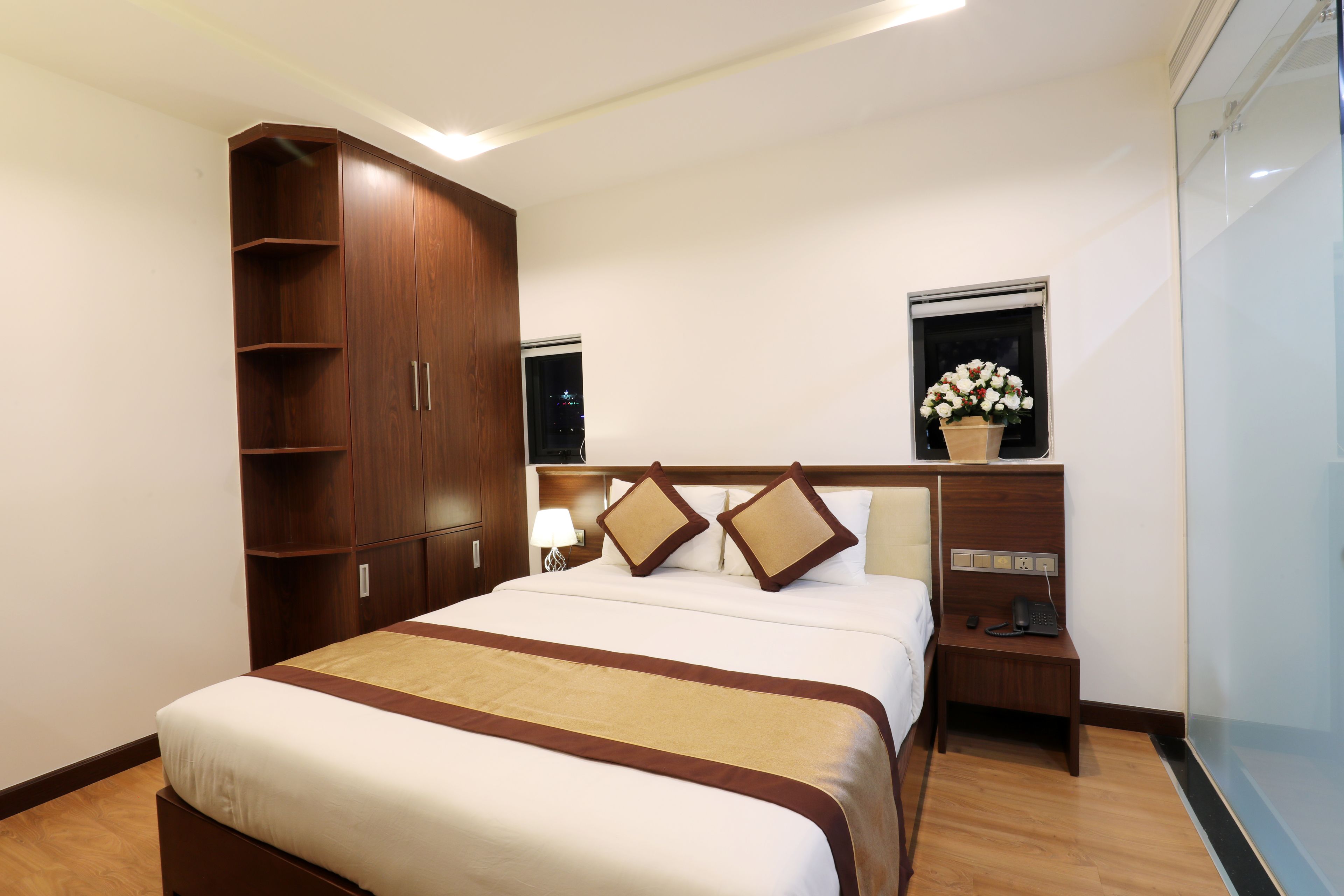 Una habitación de hotel con una cama grande, armario de madera, mesita de noche con lámpara y ventana con vistas a la ciudad de noche.