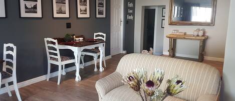A dining area with a white table and chairs, framed black-and-white photographs on the wall, a mirror above the table, and a sofa with a vase of flowers in the foreground.