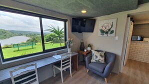 A room with a desk, a chair, a sofa, a TV mounted on the wall, and a view of a lake through a large window. - Riverview Guesthouse (East London)