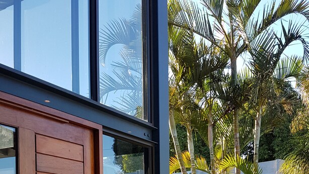 A modern building with large glass windows, a wooden door, and a stone wall with palm trees in the background. - Riverview Guesthouse (East London)