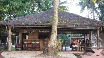 Eine rustikale Holzkonstruktion mit einem Strohdach, beschriftet mit "The Whale Bar", mit Barbereich, Sitzgelegenheiten und einem großen Baum im Vordergrund.