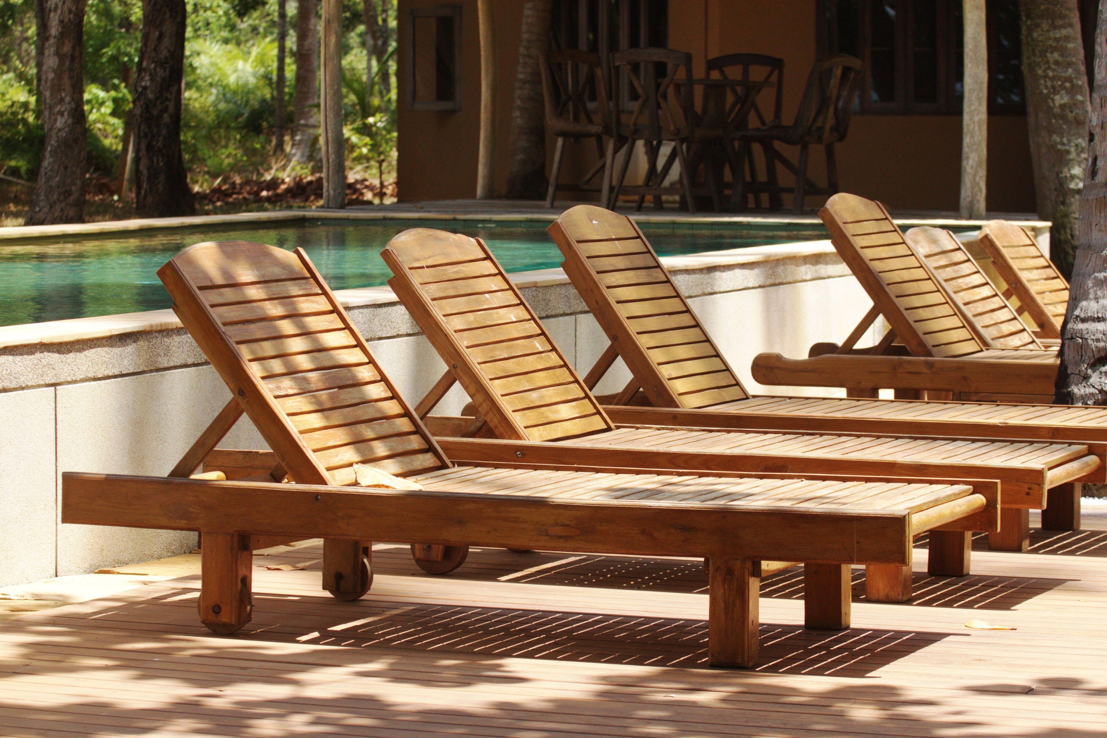 Wooden lounge chairs by a poolside with a view of trees and a building in the background.