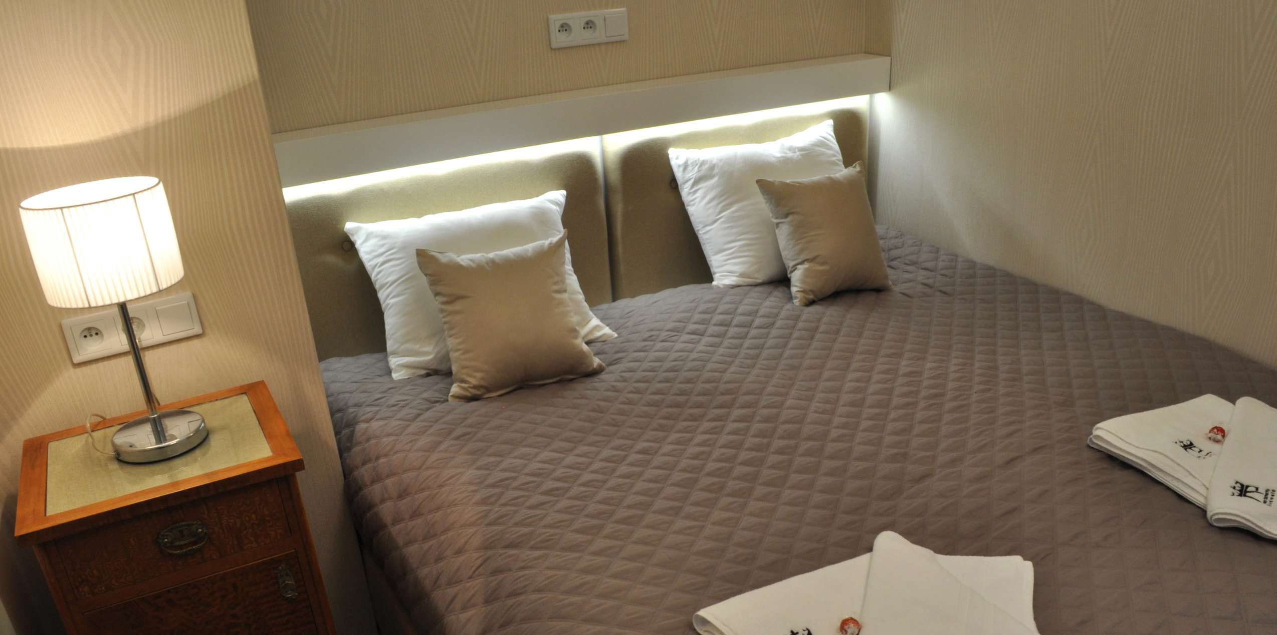Ein ordentlich bezogenes Bett mit einer gesteppten Decke, zwei Kissen, einer Nachttischlampe und einem kleinen Holennachttisch.