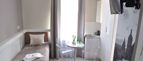 A compact hotel room with a single bed, a small kitchenette, a dining area with a table and chairs, and a large window with curtains.