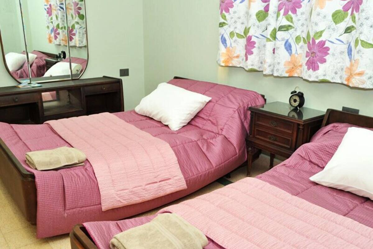 Una habitación con dos camas, cada una con ropa de cama rosa y almohadas blancas, una mesita de noche de madera con un reloj y un espejo en la pared.
