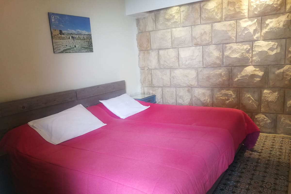 Una cama con colcha roja y almohadas blancas, pared de piedra y un cuadro enmarcado.