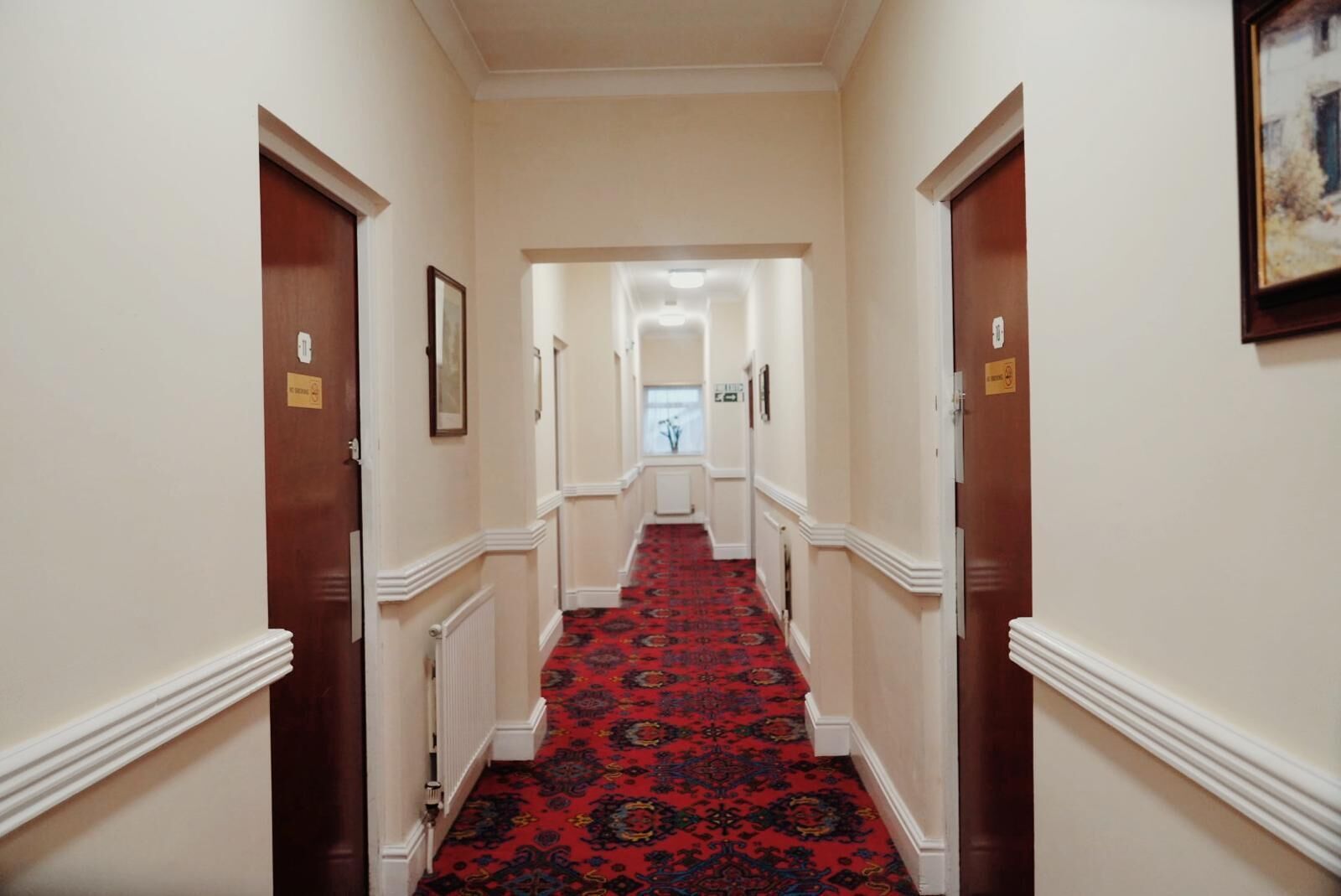 Un couloir avec un tapis à motifs rouges, des murs de couleur crème et des portes en bois.