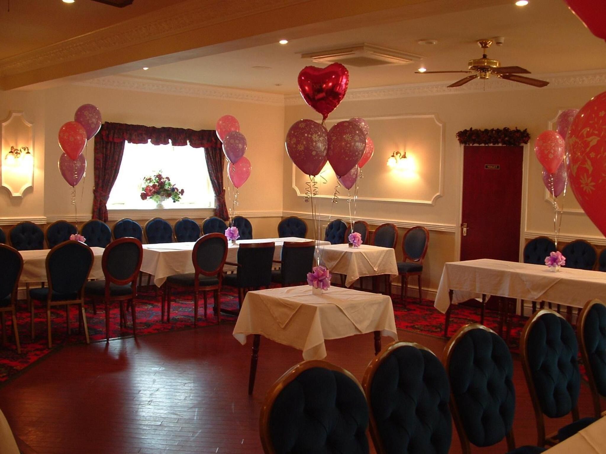 Un salón de banquetes con mesas redondas y sillas, globos y un ventanal con cortinas.