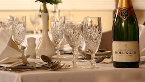 Uma mesa posta com taças de cristal, uma garrafa de champanhe Bollinger e um vaso com flores vermelhas.