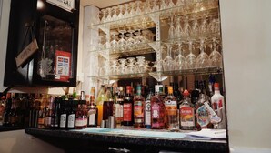 Uma área de bar com resfriador de vinhos, prateleiras com copos de vinho e diversas garrafas de bebidas alcoólicas.