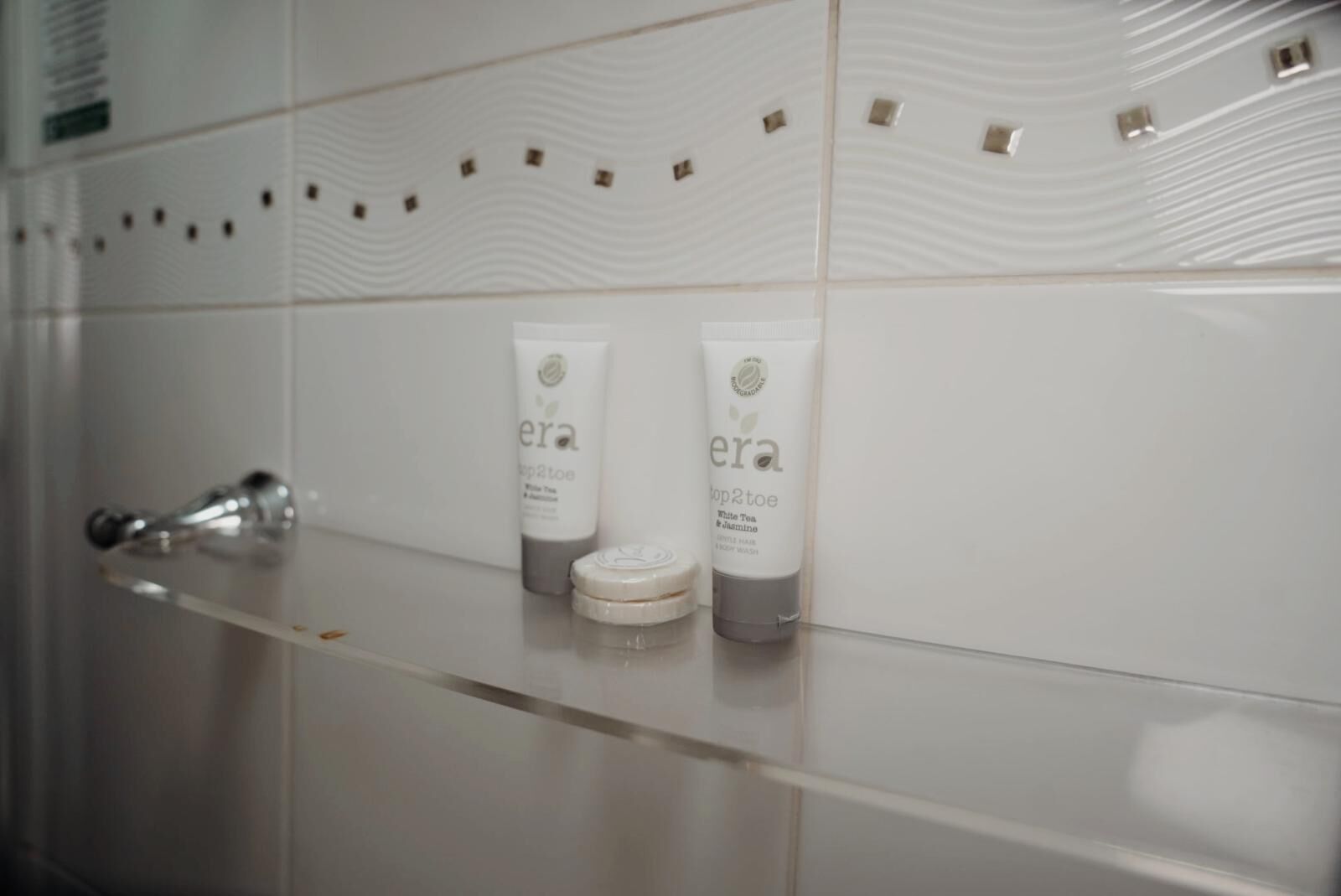 A bathroom with a white tiled wall, a glass shelf holding two bottles of 'era' products, and a silver faucet.