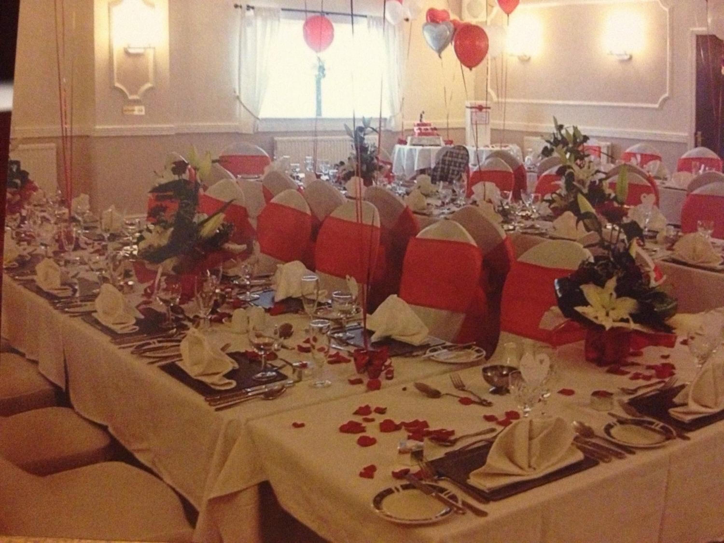 Une salle de banquet avec des tables rondes dressées pour un événement formel, agrémentée de housses de chaise rouges et blanches, de compositions florales et de pétales de roses disposés sur les tables.