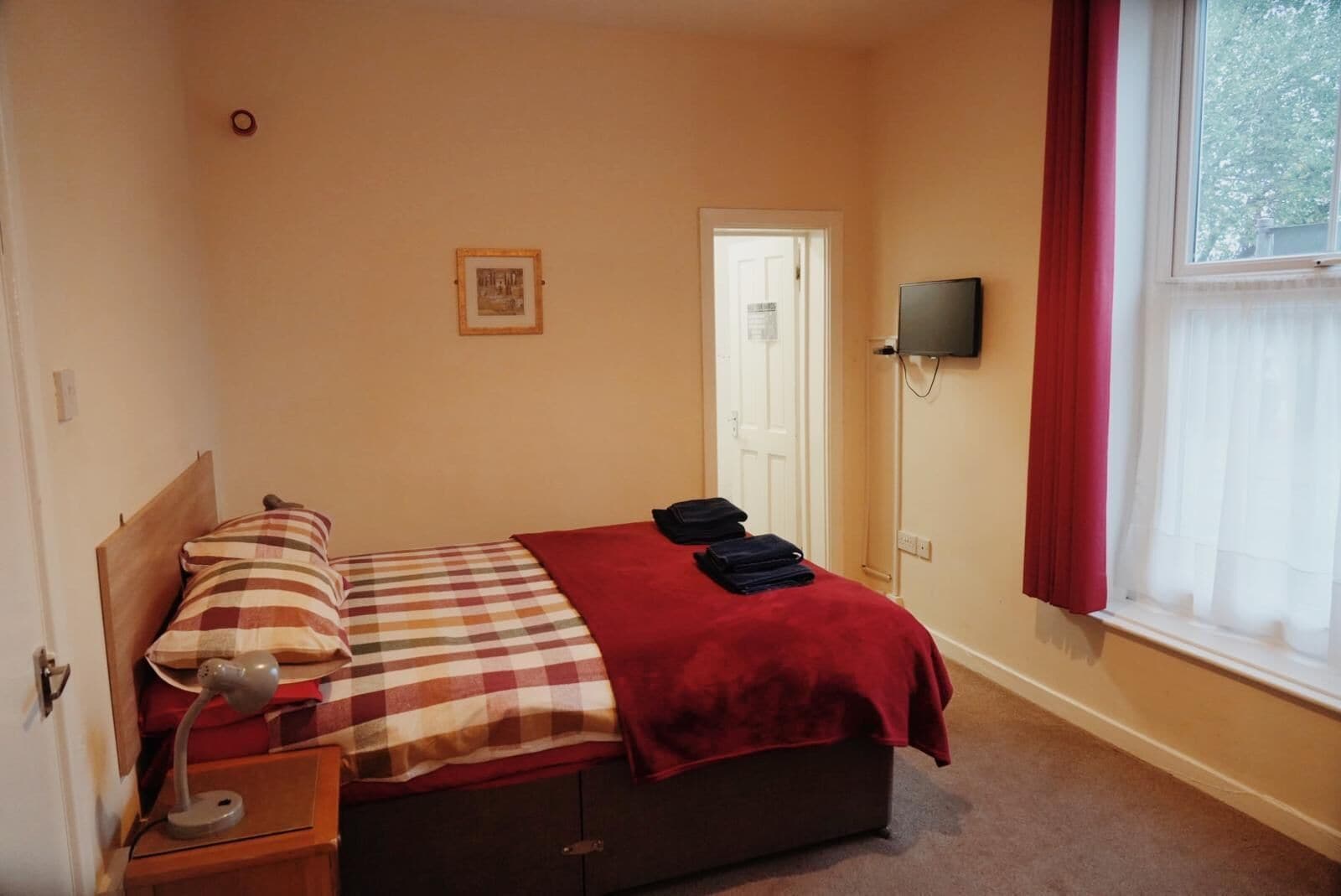 Un dormitorio con cama, mesita de noche, lámpara, televisor y ventana con cortinas.