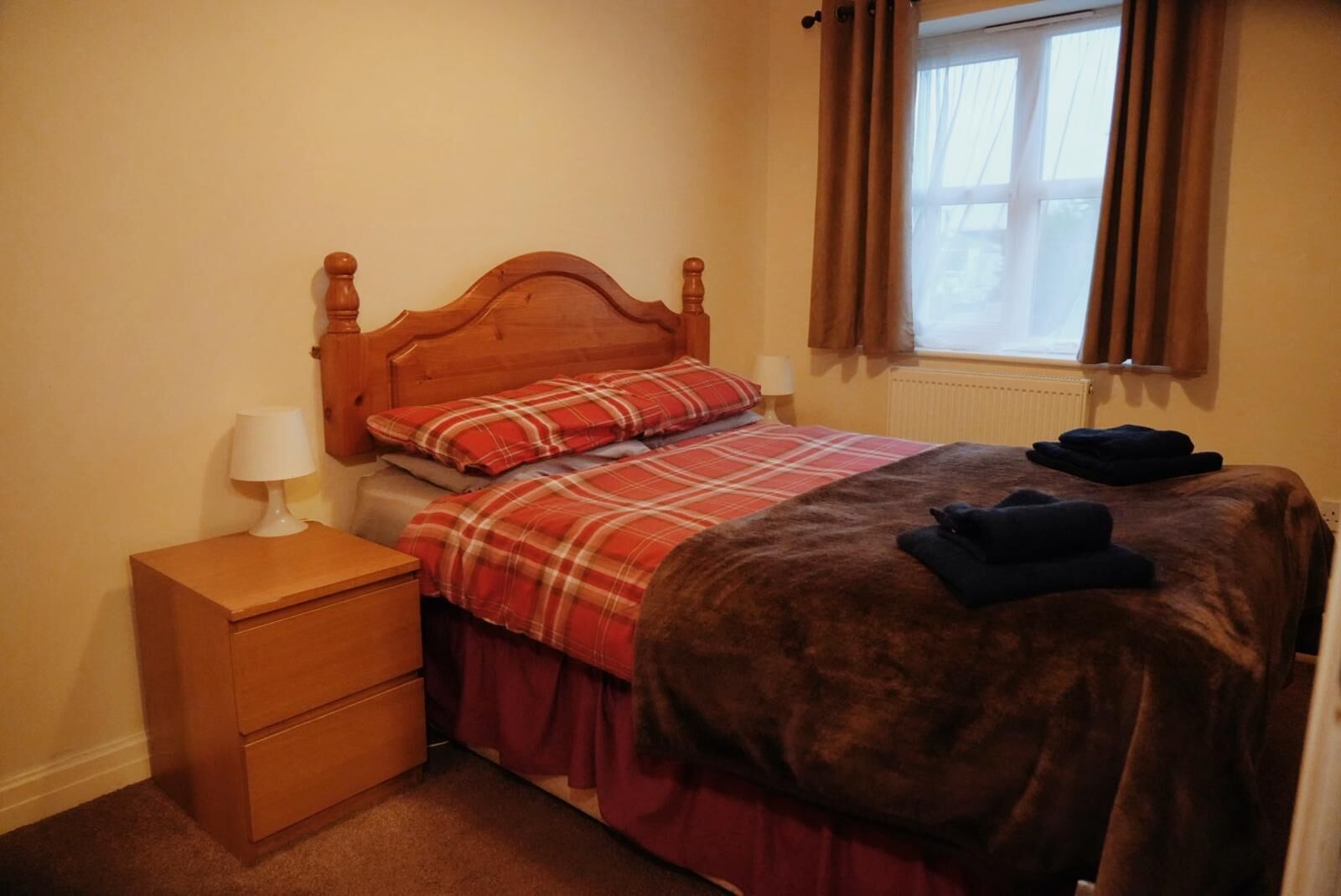 Um quarto pequeno e bem organizado, com uma cama de madeira, uma mesinha de cabeceira com abajur e uma janela com cortinas.