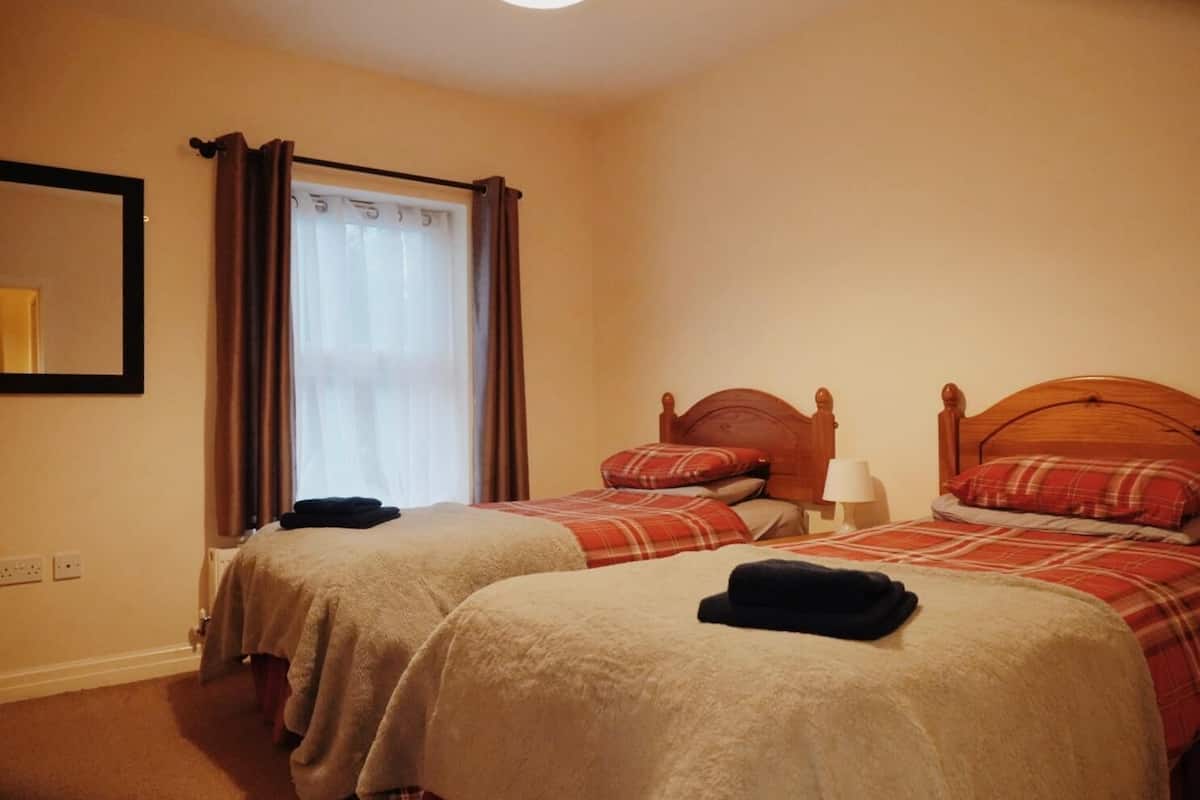 A hotel room with two beds, each with a wooden headboard, a nightstand with a lamp, and a window with curtains.