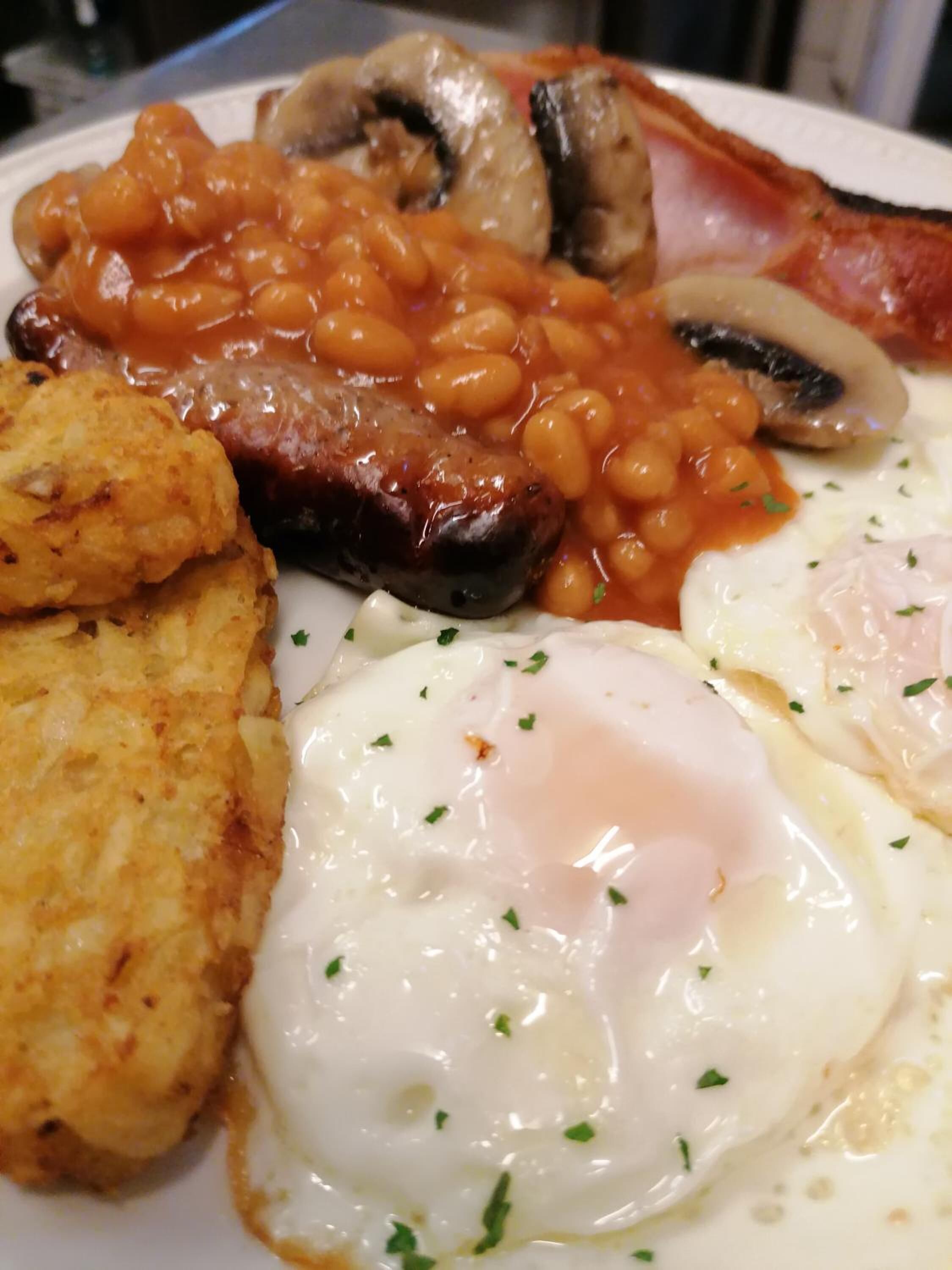 A plate of breakfast food including beans, bacon, mushrooms, hash browns, and fried eggs.