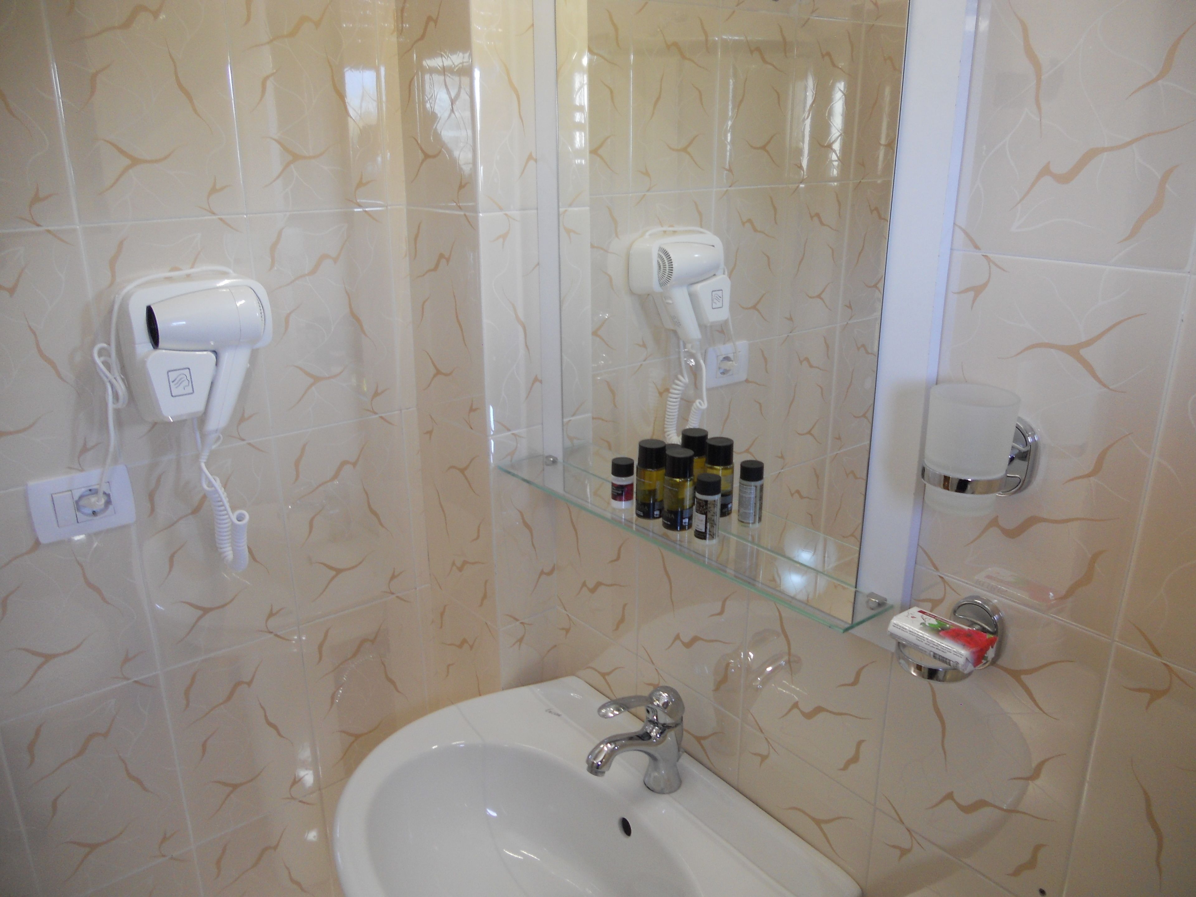 Uma casa de banho com uma pia branca, um secador de cabelo e uma prateleira de vidro com artigos de higiene pessoal.