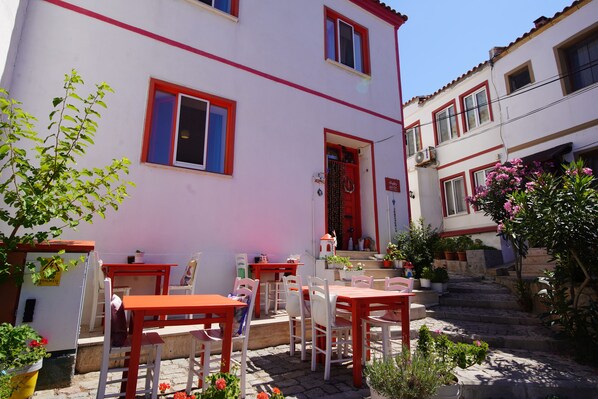 A building with a red door and windows, outdoor seating with white chairs and tables, and potted plants. - Alaybey Ada Evi (Bozcaada)