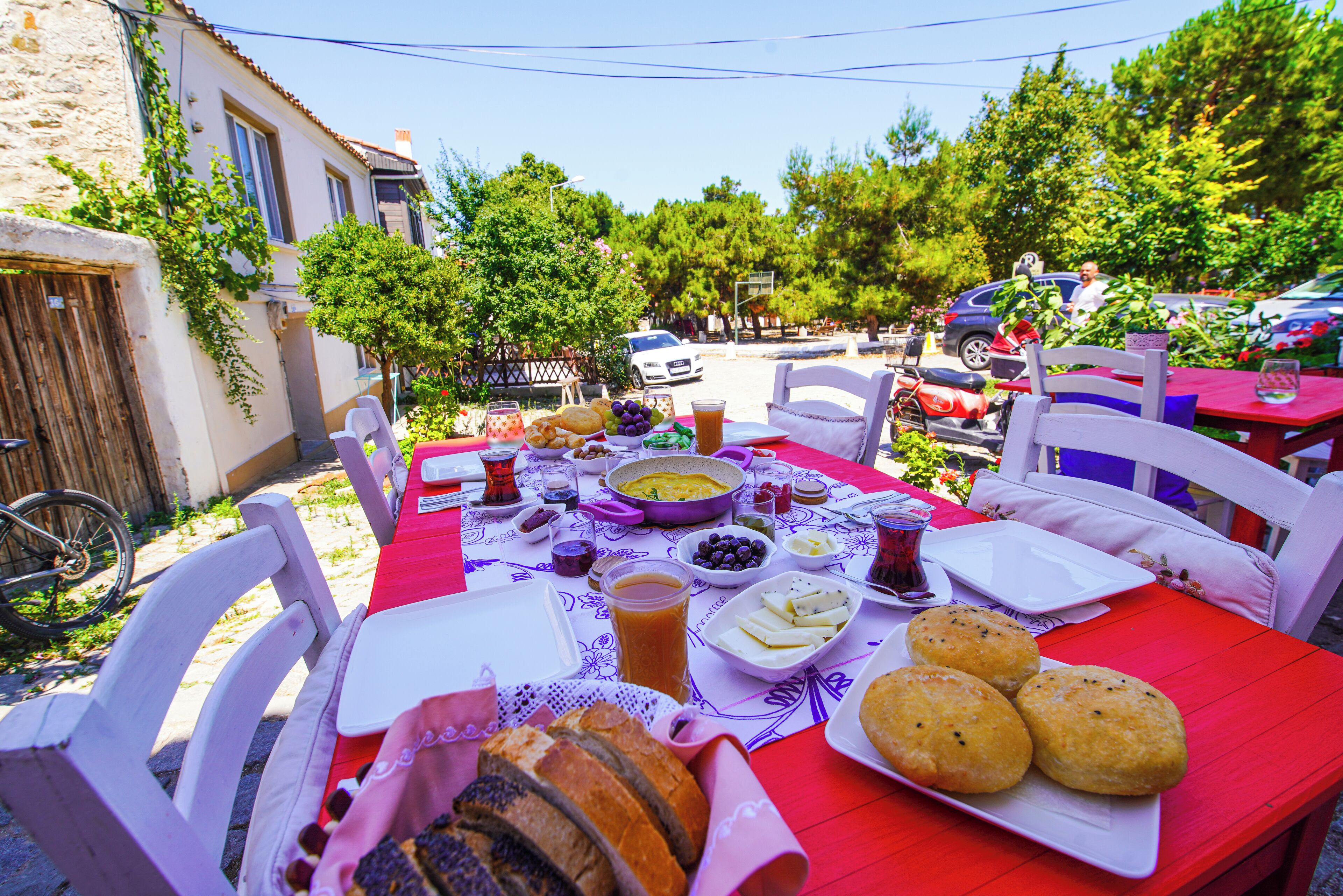An outdoor dining table with food and drinks, surrounded by white chairs, in a sunny outdoor setting.