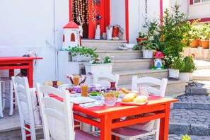 An outdoor dining area with a red table and white chairs, surrounded by potted plants and a white building with red accents. - Alaybey Ada Evi (Bozcaada)