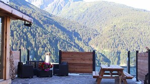 An outdoor seating area with a wooden bench and table, surrounded by stone flooring, with a mountain view in the background. - Hotel Riva Sport & Wellness (Vione)