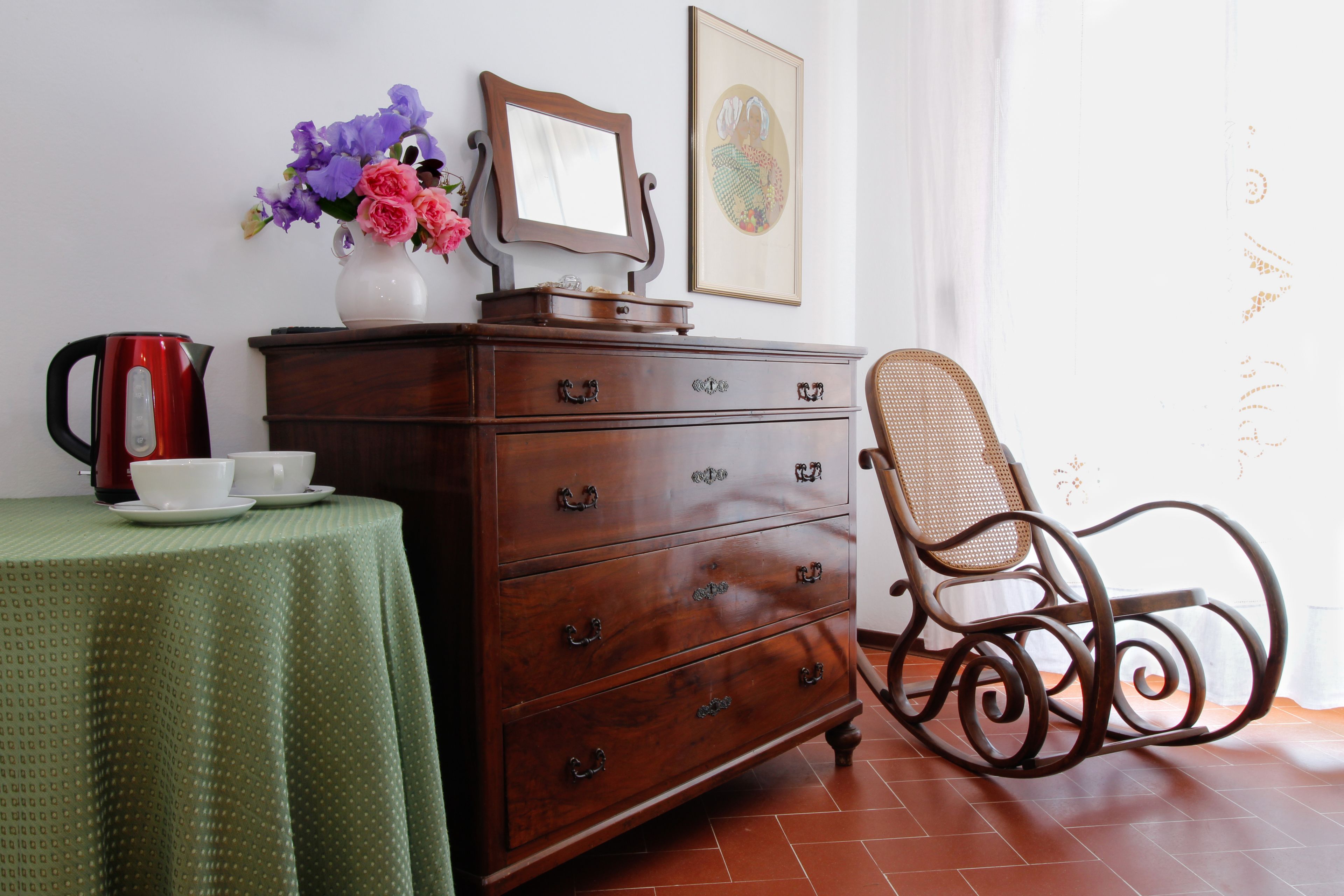 A wooden dresser with drawers, a mirror, and a vase with flowers. There is a chair with a woven seat and a table with a green tablecloth and a teapot.