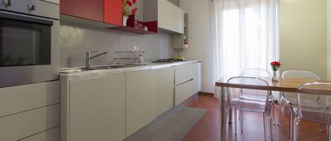 A modern kitchen with white cabinets, a stainless steel oven, and a red accent wall. There is a dining area with a wooden table and metal chairs.