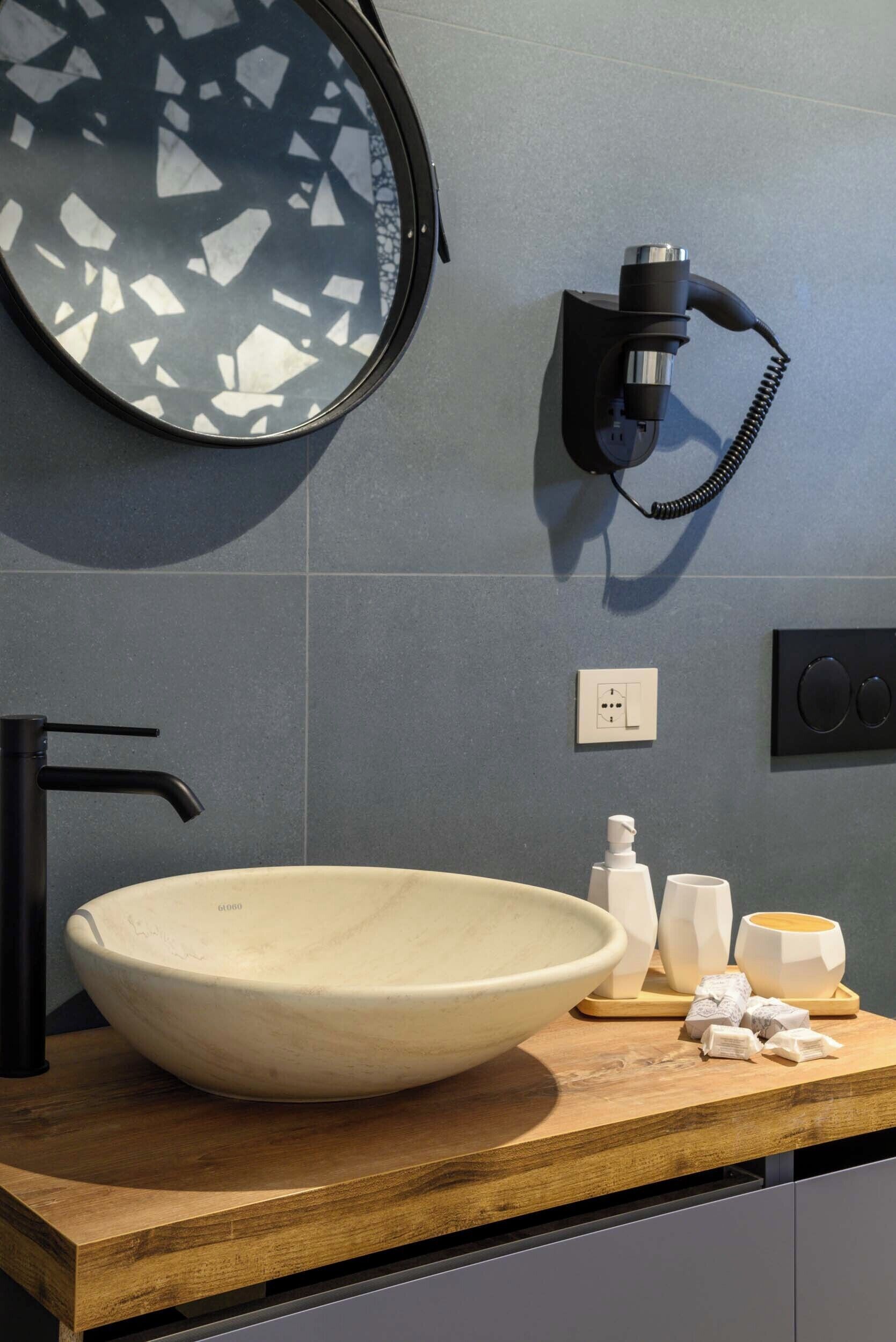 A modern bathroom with a large, round, beige sink, a black faucet, a wall-mounted hairdryer, and a mirror with a geometric pattern.