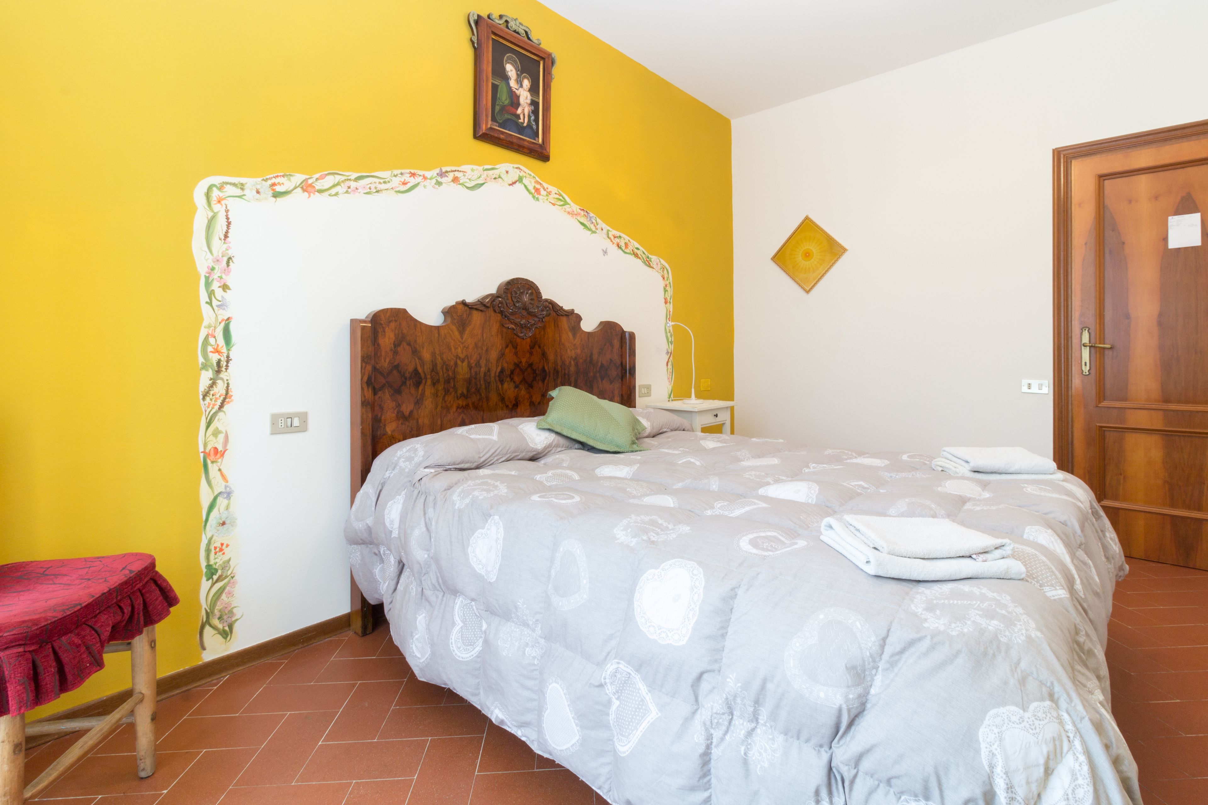 A bedroom with a wooden headboard, a bed with a grey patterned bedspread, a green pillow, and a red bench.
