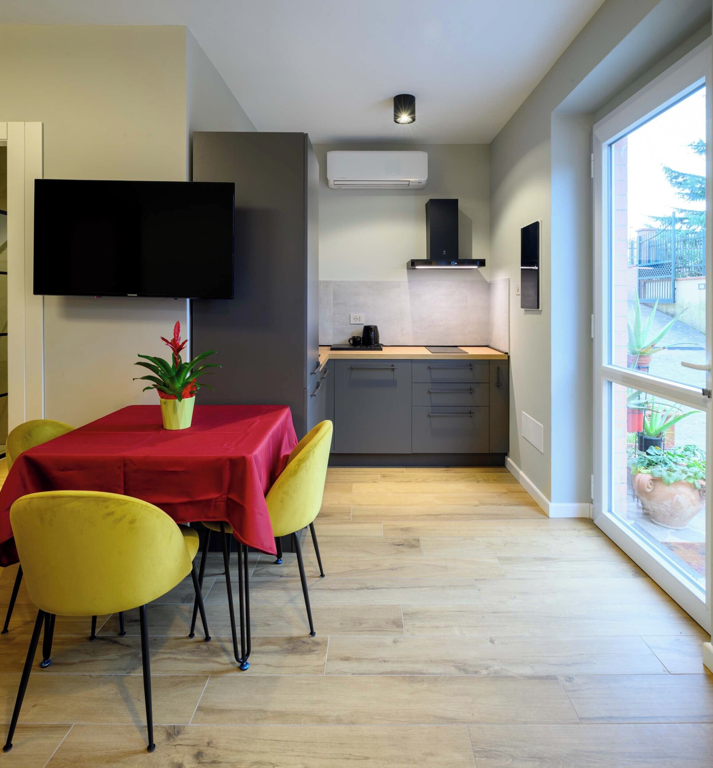 Una cocina moderna con un televisor de pantalla plana, un comedor con una mesa roja y sillas amarillas, y un ventanal con vistas al exterior.