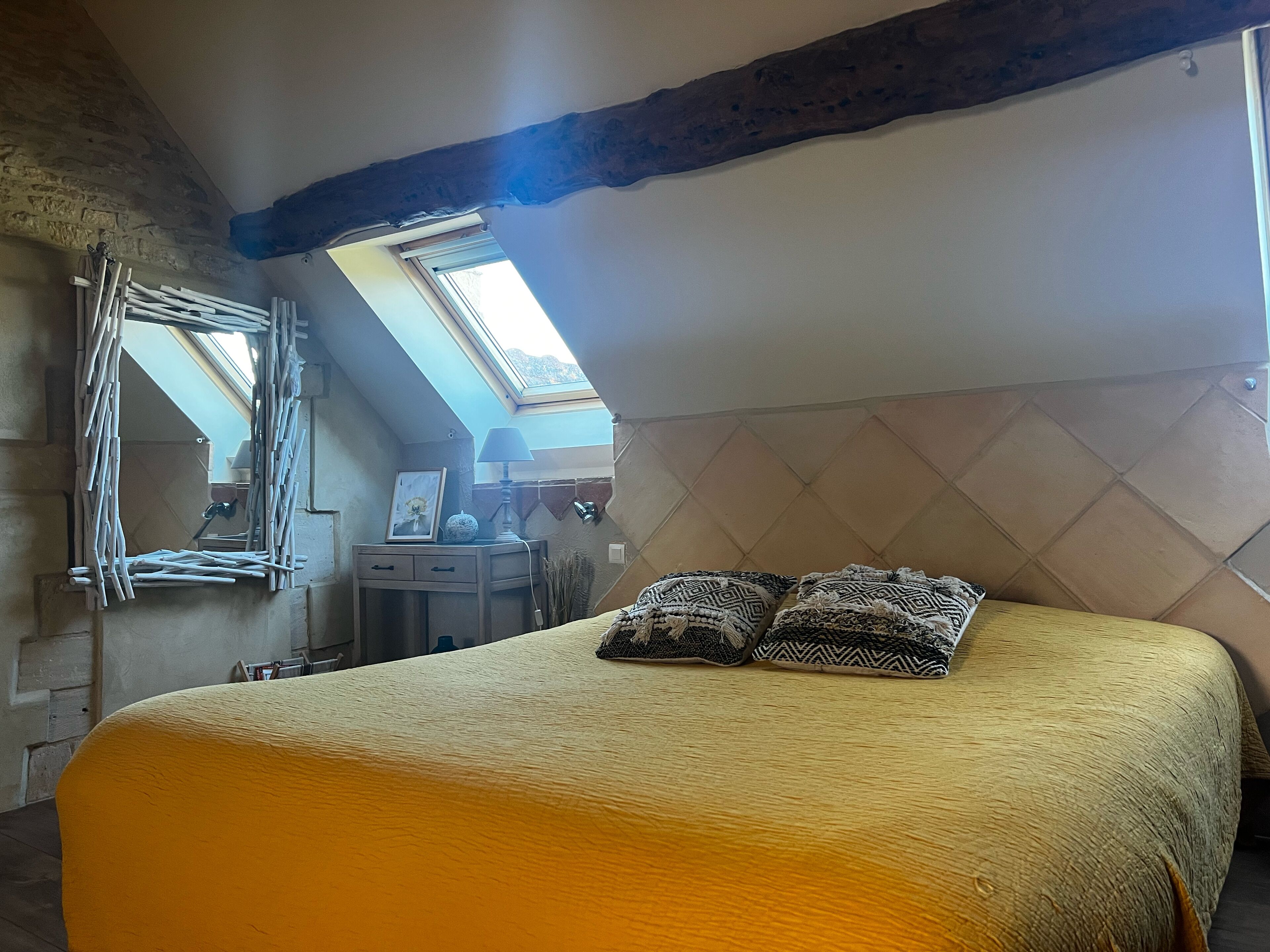 A bedroom with a sloped ceiling, a bed with patterned pillows, a desk with a lamp, and a framed picture.