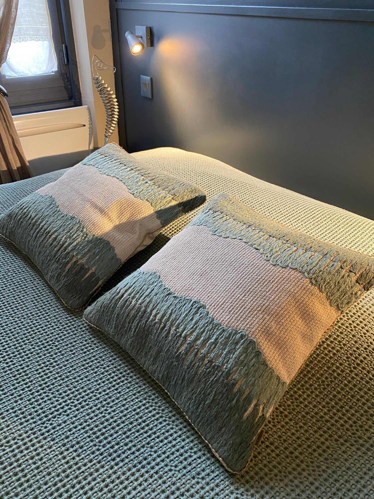 Un lit avec une couverture et des oreillers à motifs, une fenêtre avec des rideaux et un luminaire fixé au mur.