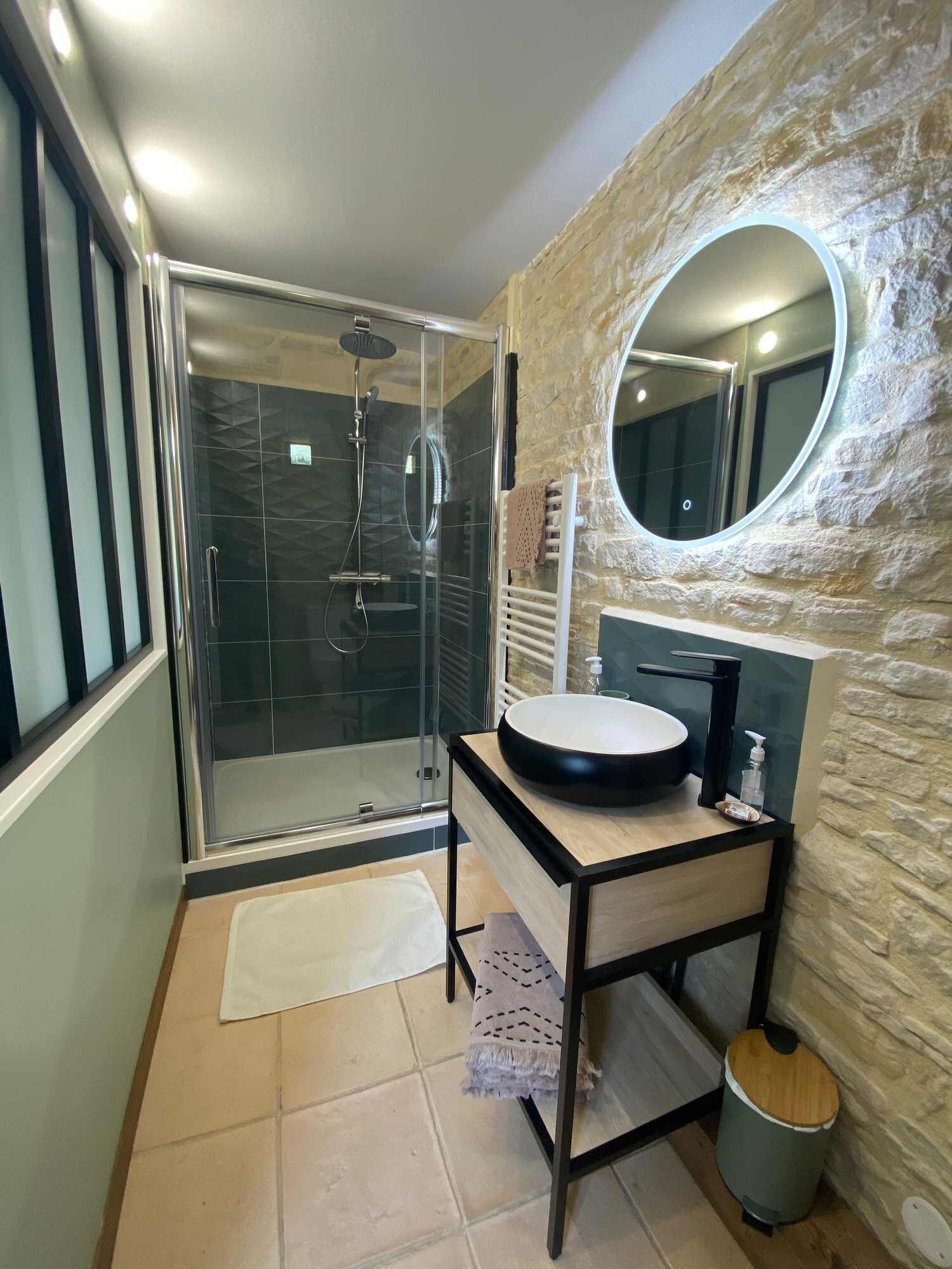 Ein modernes Badezimmer mit einer gläsernen Duschkabine, einem runden Waschbecken mit schwarzer Arbeitsplatte und einer Wand mit Steinoptik.