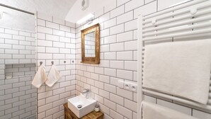 Un baño con azulejos blancos tipo metro, un tocador de madera, un lavamanos blanco, un espejo con marco dorado y un perchero con dos toallas.