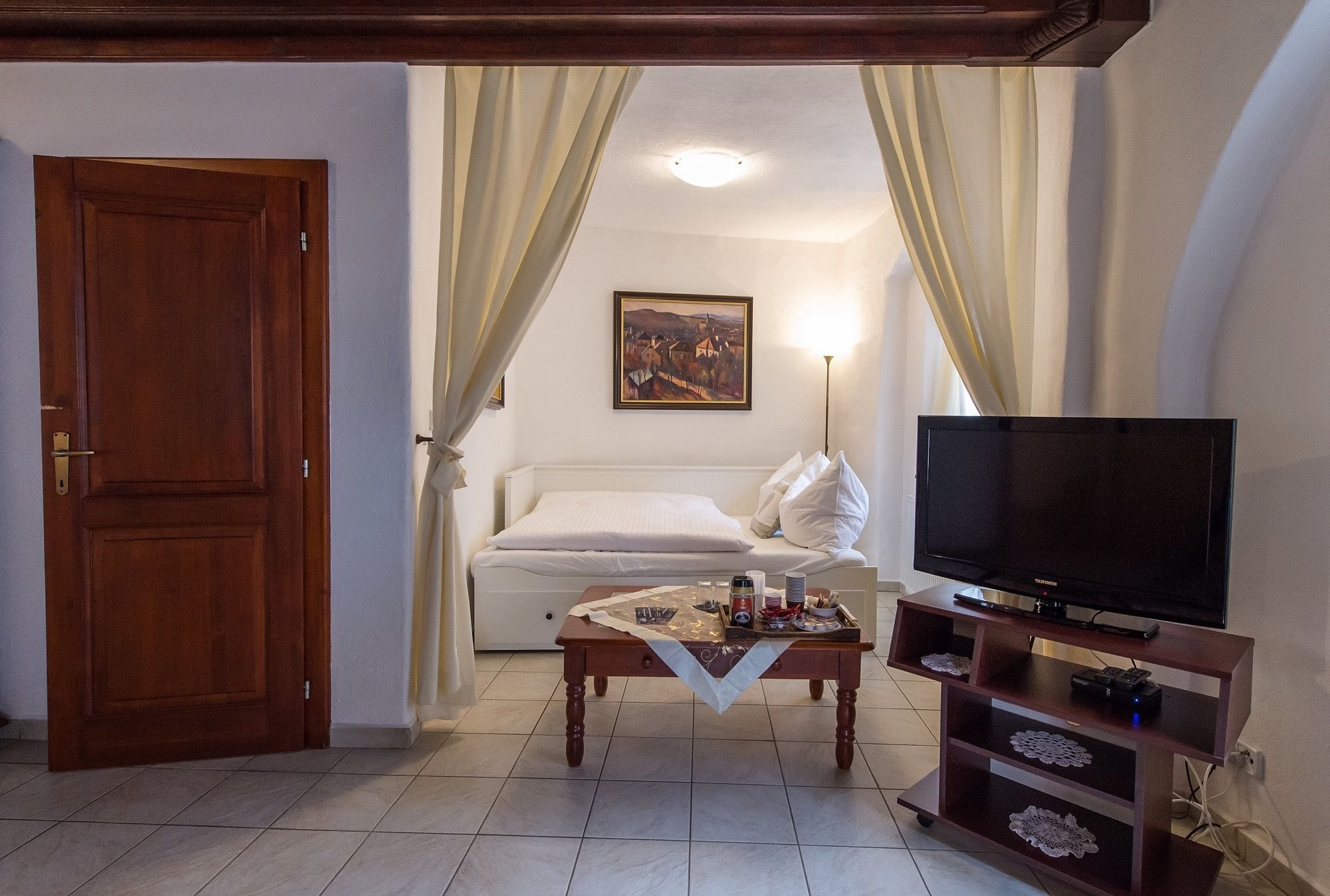 Una habitación con puerta de madera, un sofá blanco, una mesita de centro, un televisor sobre un mueble de madera y un cuadro en la pared.