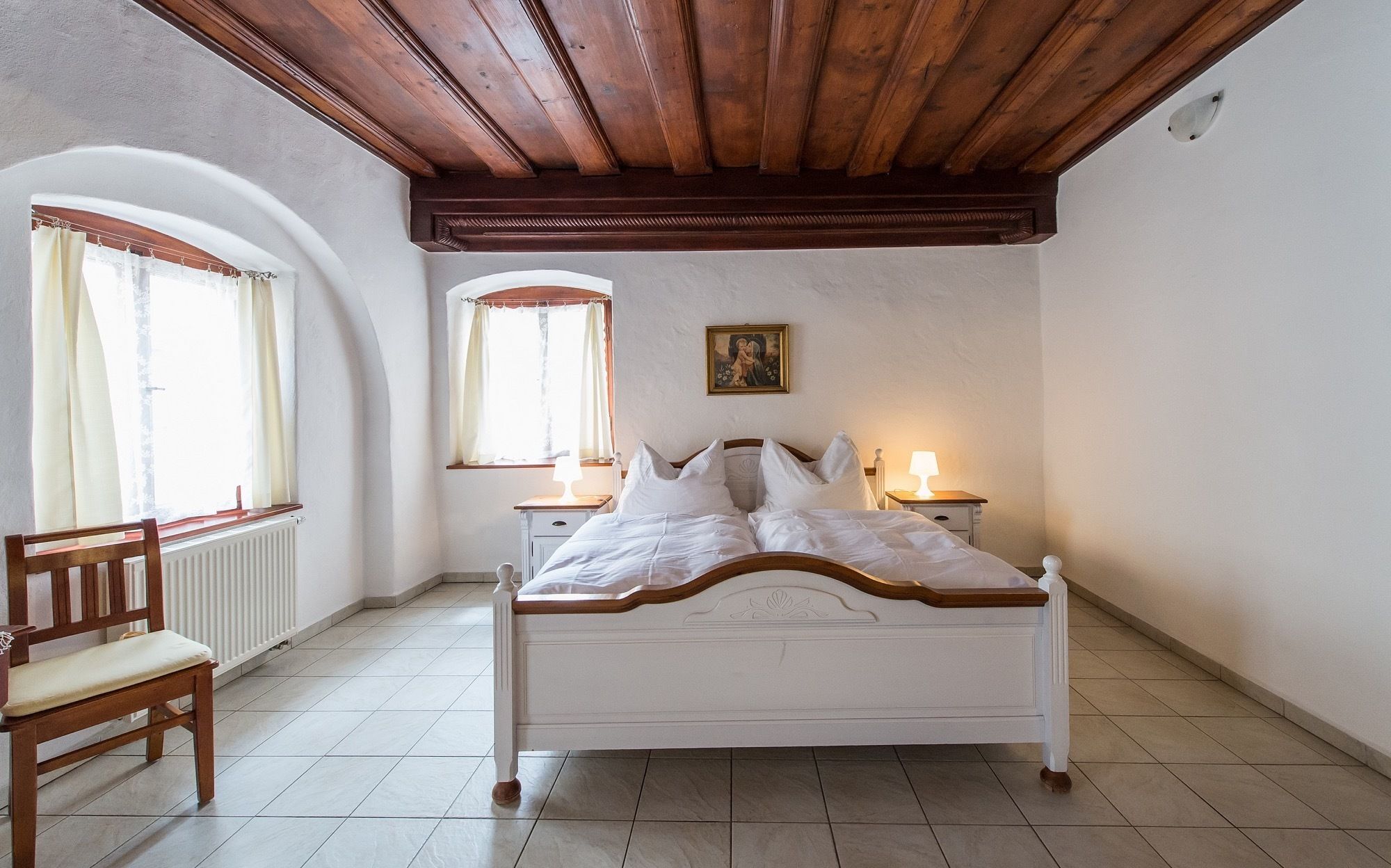 Een slaapkamer met een houten bed, twee nachtkastjes met lampen, een stoel en een raam met gordijnen.