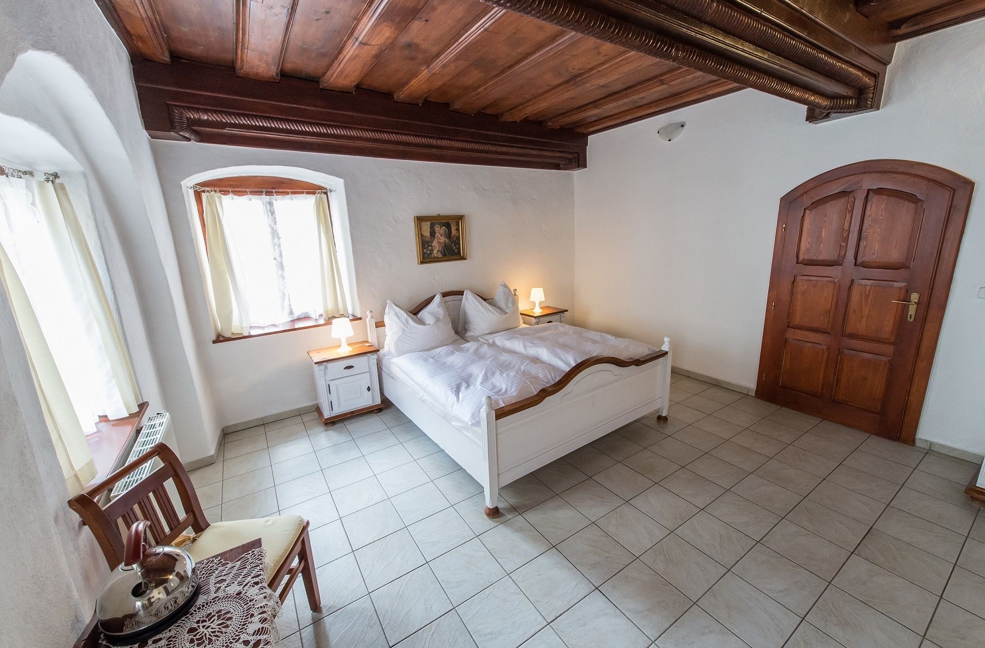 Una camera da letto con un letto, un comodino, una finestra con tende, una porta in legno e una sedia.