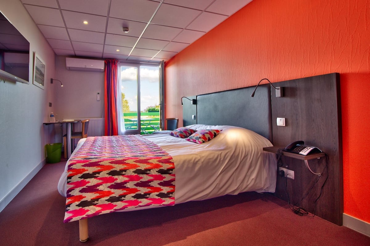 A modern hotel room with a large bed, a desk, and a view of a green outdoor area.