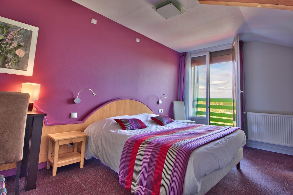 A bedroom with a bed, bedside tables, a nightstand, a lamp, a painting, and a window with a view.