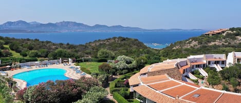 A resort with a swimming pool surrounded by sun loungers, a well-maintained garden, and a view of the sea and mountains.