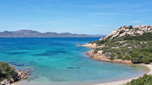 Eine Küstenlandschaft mit klarem türkisblauem Wasser, einem Sandstrand, felsigem Gelände und Bergen in der Ferne.