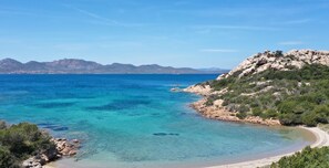 Eine Küstenlandschaft mit klarem türkisblauem Wasser, einem Sandstrand, felsigem Gelände und Bergen in der Ferne.