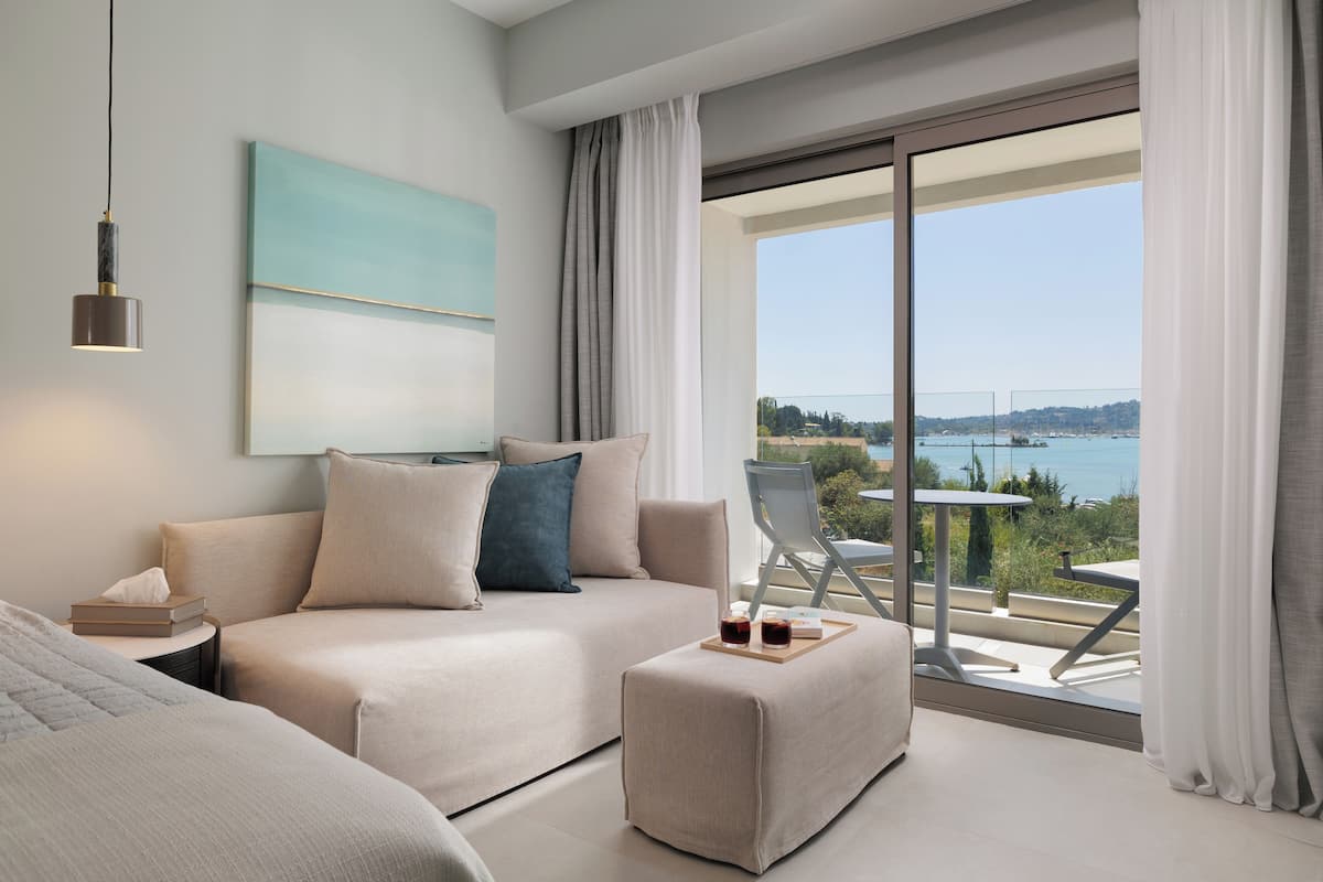 A modern bedroom with a large window, a sofa, a small table with drinks, and a painting on the wall.