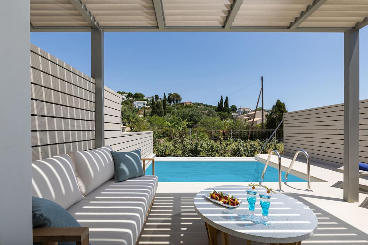 A poolside area with a sofa, a table with drinks, and a view of a swimming pool and greenery.