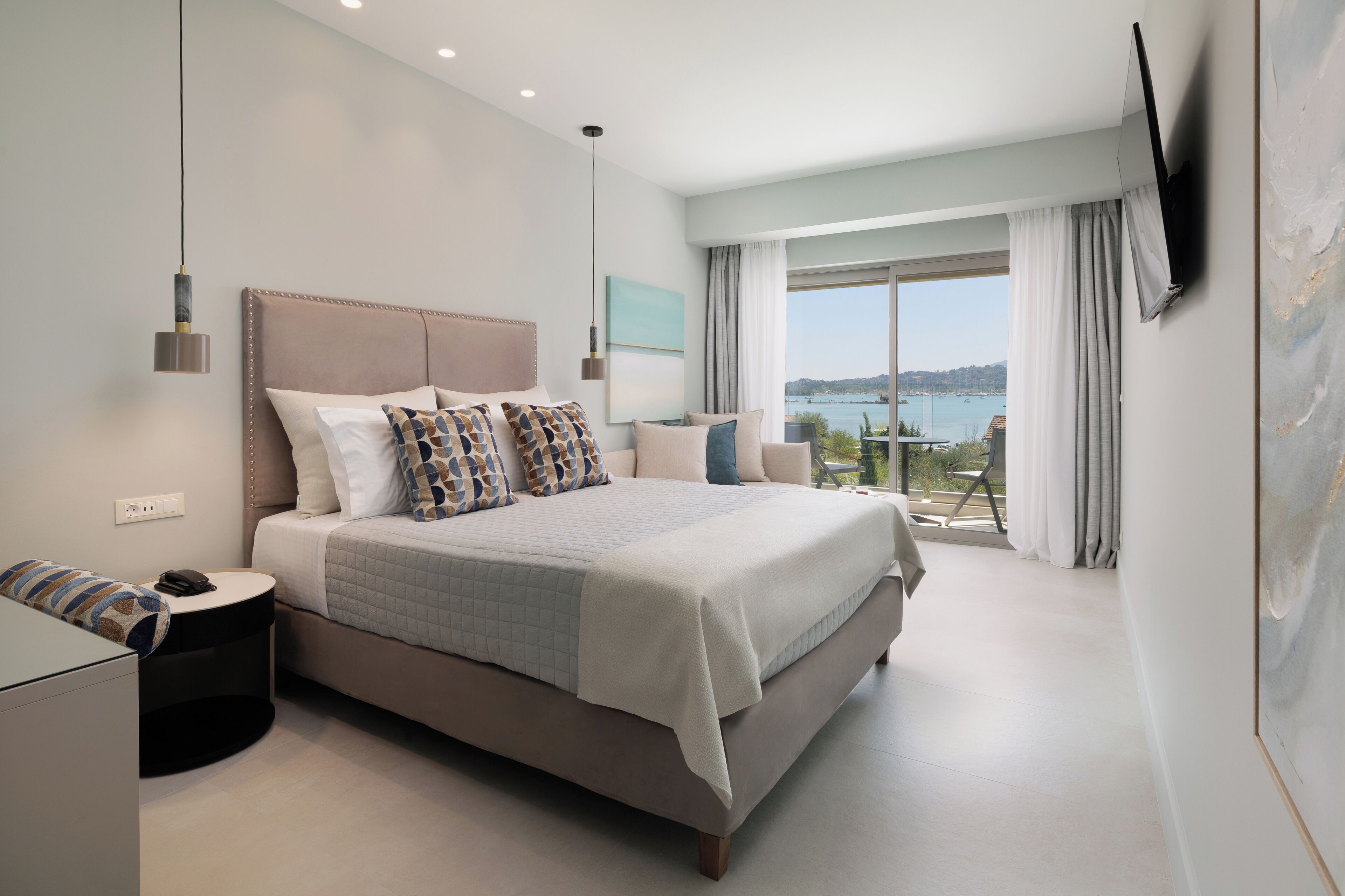 A modern bedroom with a large bed, a bedside table, a TV mounted on the wall, and a view of the sea through large windows.