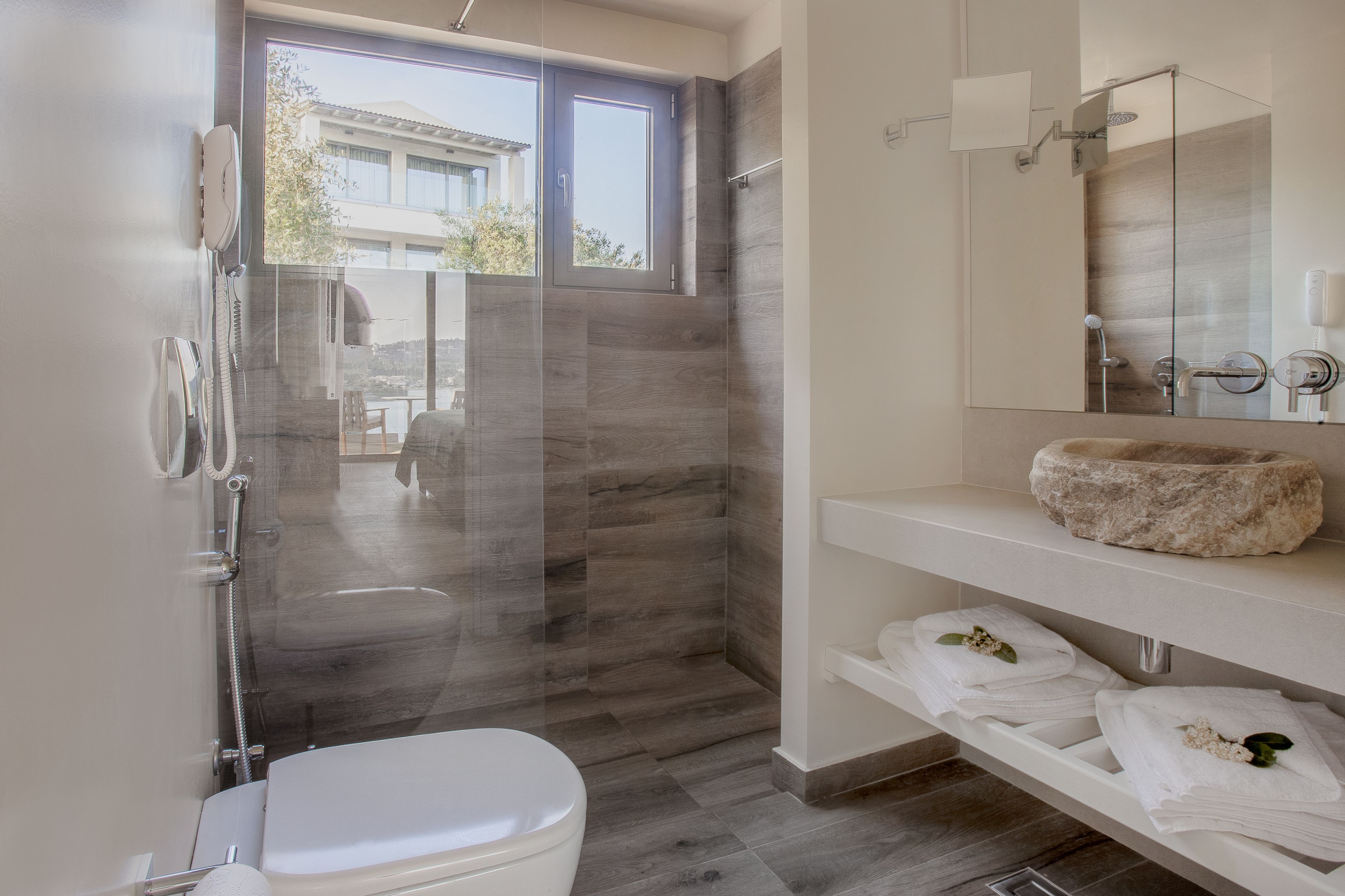 Un baño moderno con ducha envuelta en vidrio, inodoro blanco, lavamanos de piedra y toallas dobladas ordenadamente en un estante.